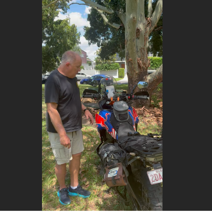 nomad moto Man standing next to a red and blue ATV in a grassy area with trees and cars in the background.