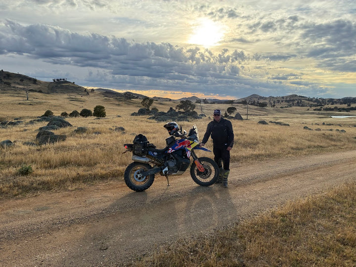 Person standing next to a motorcycle on a dirt road with a scenic landscape and sunset.
